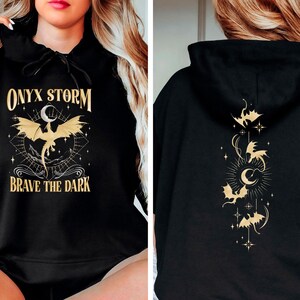 May include: Black hoodie with a gold graphic of a dragon and the text "ONYX STORM" and "BRAVE THE DARK". The back of the hoodie has a gold graphic of a crescent moon and several dragons.