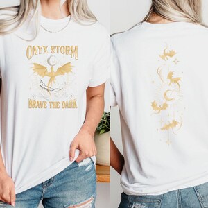 May include: White t-shirt with a gold graphic of a dragon and the text "ONYX STORM BRAVE THE DARK". The back of the shirt has a gold graphic of multiple dragons.