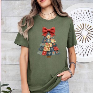 May include: An olive green t-shirt with a Christmas tree design. The tree is made of layered fabric pieces in various patterns and colours, topped with a red bow. The design includes floral and snowflake patterns.