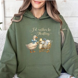 May include: An olive green hoodie with a graphic of a duck wearing a blue bow tie, pulling a shopping trolley filled with vintage items. The text above the duck reads "I'd rather be Thrilling".