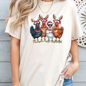 May include: A cream-coloured t-shirt with a graphic of five chickens in Christmas attire. Each chicken is wearing a Santa hat and reindeer antlers. Some are decorated with fairy lights and scarves, creating a festive holiday design.
