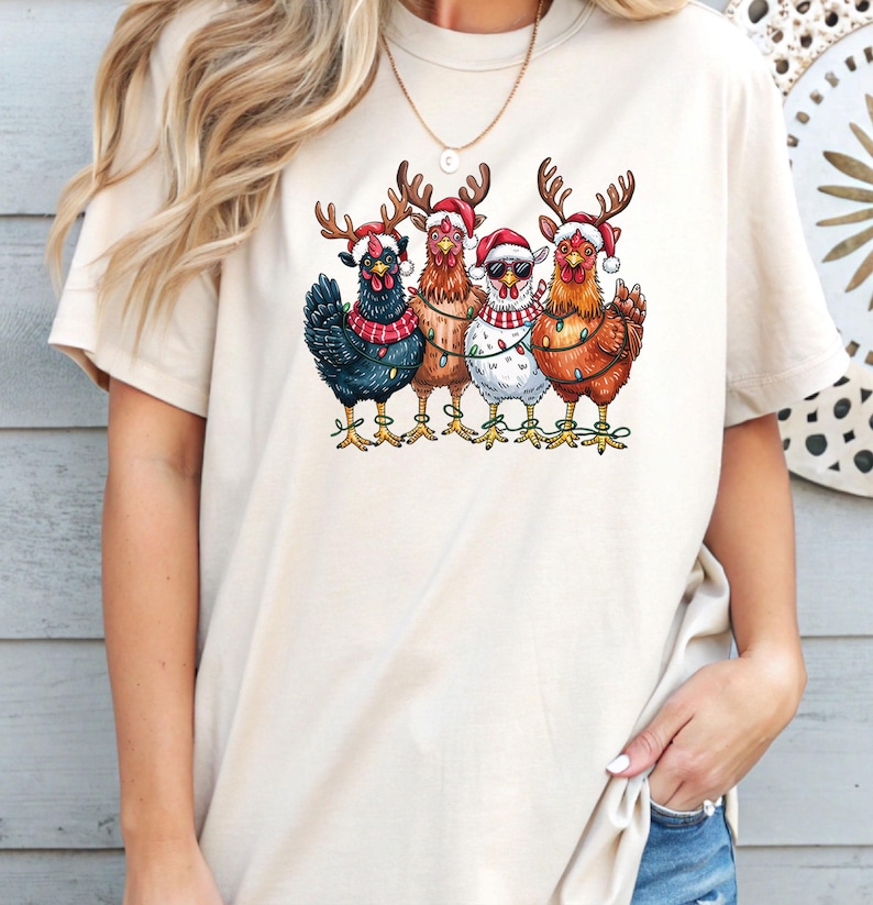 May include: A cream-coloured t-shirt with a graphic of five chickens in Christmas attire. Each chicken is wearing a Santa hat and reindeer antlers. Some are decorated with fairy lights and scarves, creating a festive holiday design.
