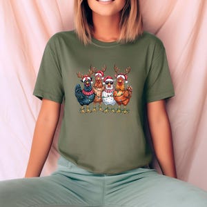May include: An olive green t-shirt with a graphic of five chickens adorned with reindeer antlers and Santa hats. The chickens are in different colours, with one wearing sunglasses and another a string of lights. A festive holiday design.