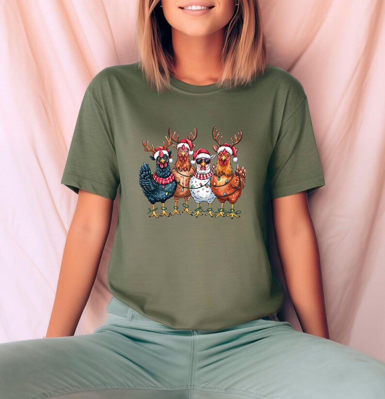 May include: An olive green t-shirt with a graphic of five chickens adorned with reindeer antlers and Santa hats. The chickens are in different colours, with one wearing sunglasses and another a string of lights. A festive holiday design.