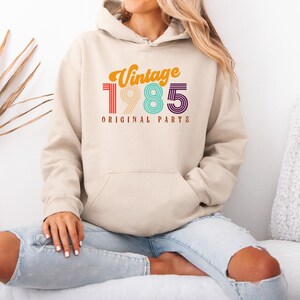 May include: A beige hoodie with a vintage 1985 design in rainbow colors. The text "Vintage 1985 Original Parts" is printed on the front of the hoodie.