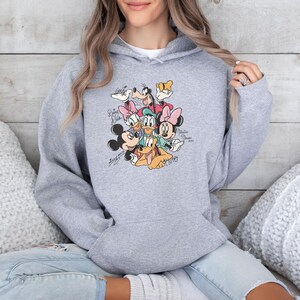 May include: A light gray hooded sweatshirt featuring a colorful graphic of Disney characters, including Mickey Mouse, Minnie Mouse, Donald Duck, and Goofy. The graphic includes character signatures. The sweatshirt is made of a soft material.