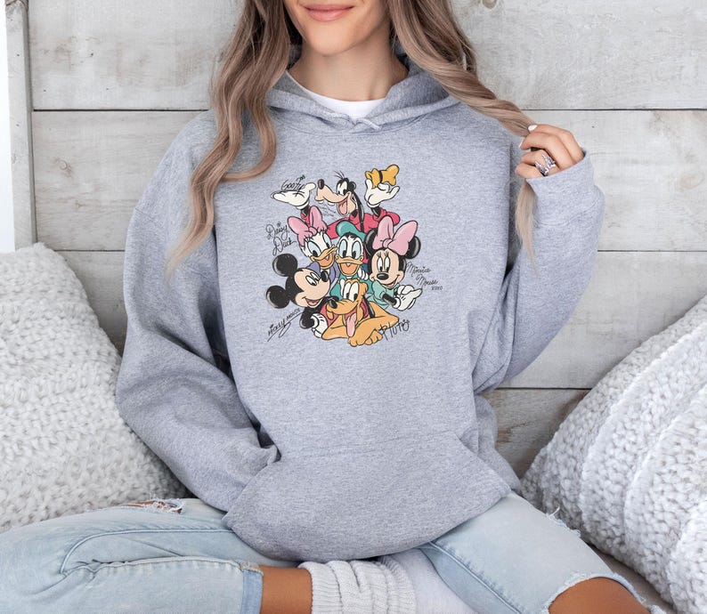 May include: A light gray hooded sweatshirt featuring a colorful graphic of Disney characters, including Mickey Mouse, Minnie Mouse, Donald Duck, and Goofy. The graphic includes character signatures. The sweatshirt is made of a soft material.
