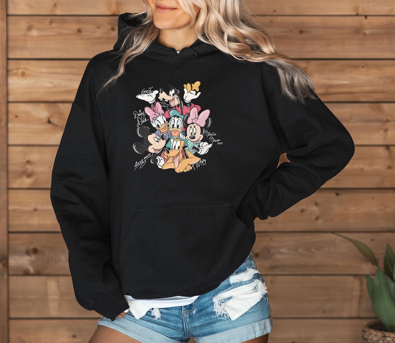 May include: Black hooded sweatshirt featuring a colorful graphic of Disney characters including Mickey Mouse, Minnie Mouse, Donald Duck, Daisy Duck, Goofy, and Pluto. The graphic includes character signatures. The sweatshirt has a front pocket.