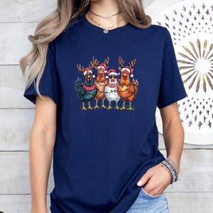 May include: A navy blue t-shirt with a Christmas-themed graphic. The design shows five chickens, each wearing a Santa hat and reindeer antlers. Some chickens are decorated with fairy lights and sunglasses. The chickens are in different colors, including black, brown, and white.