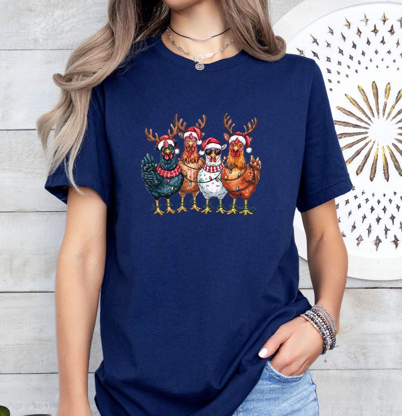 May include: A navy blue t-shirt with a Christmas-themed graphic. The design shows five chickens, each wearing a Santa hat and reindeer antlers. Some chickens are decorated with fairy lights and sunglasses. The chickens are in different colors, including black, brown, and white.