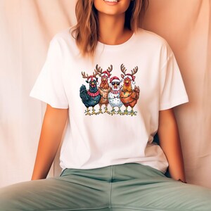 May include: White t-shirt with a Christmas-themed design of five chickens. Each chicken is wearing a Santa hat and reindeer antlers, with one wearing sunglasses. The chickens are decorated with string lights.