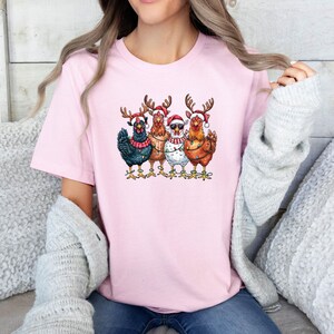 May include: A pale pink t-shirt with a festive graphic of four chickens. Each chicken is adorned with Christmas hats, reindeer antlers, and fairy lights. The chickens are depicted in shades of black, brown, and white.