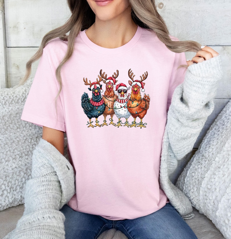 May include: A pale pink t-shirt with a festive graphic of four chickens. Each chicken is adorned with Christmas hats, reindeer antlers, and fairy lights. The chickens are depicted in shades of black, brown, and white.