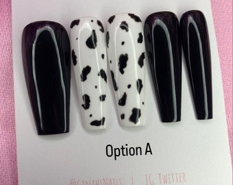 May include: A set of five artificial nails in a coffin shape. The nails are a mix of solid black and white with black cow print designs. The text "Option A" is below the nails. The brand name "GinahsNails" is printed on the card.