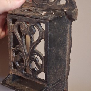 May include: Ornate cast iron match holder with a decorative floral design. The rectangular box has an openwork design on the sides and a small tray at the bottom. The top features a decorative crown-like element. The match holder is dark brown.