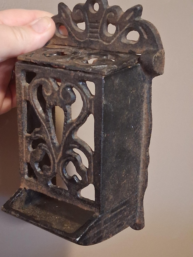 May include: Ornate cast iron match holder with a decorative floral design. The rectangular box has an openwork design on the sides and a small tray at the bottom. The top features a decorative crown-like element. The match holder is dark brown.