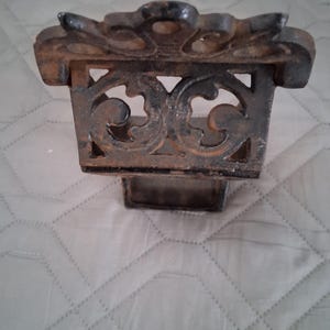 May include: Ornate, cast iron decorative piece with a rectangular base and a detailed, openwork design. The top features a stylized floral or leaf pattern. The piece has a dark, aged appearance, suggesting it may be vintage or antique.