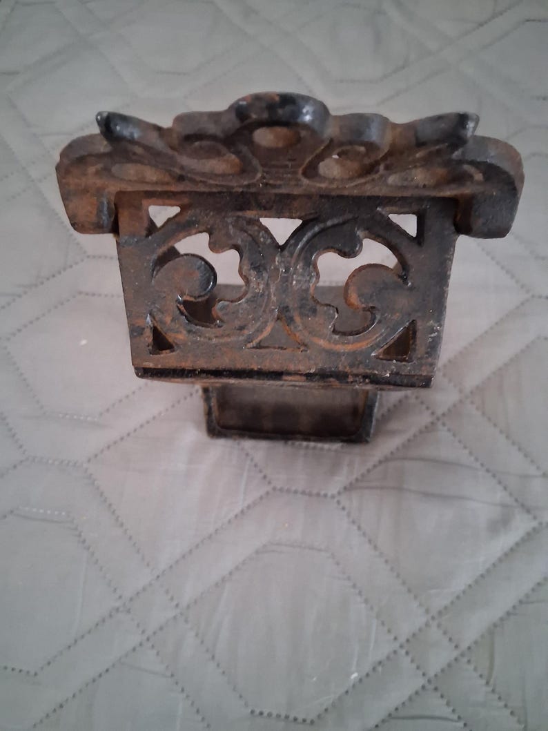 May include: Ornate, cast iron decorative piece with a rectangular base and a detailed, openwork design. The top features a stylized floral or leaf pattern. The piece has a dark, aged appearance, suggesting it may be vintage or antique.