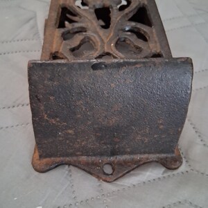 May include: A rectangular, cast iron match holder with an ornate, openwork design on the top. The match holder has a dark, rusty brown color and a small hole at the bottom. The front has a curved shape.