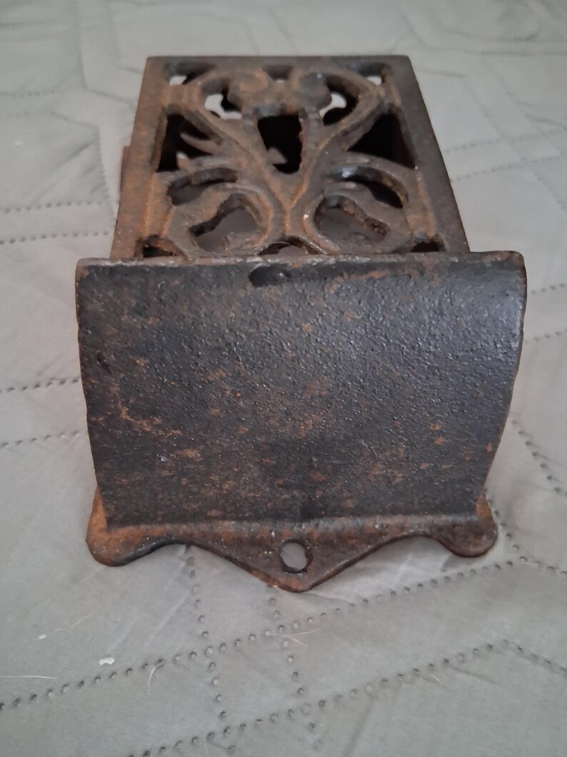 May include: A rectangular, cast iron match holder with an ornate, openwork design on the top. The match holder has a dark, rusty brown color and a small hole at the bottom. The front has a curved shape.