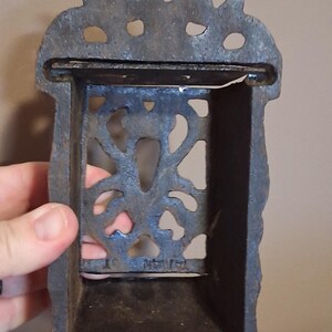 May include: A cast iron match holder with a decorative floral design. The holder is rectangular with a small shelf and a slot for matches. The metal has a dark, aged appearance, suggesting it is vintage or antique.