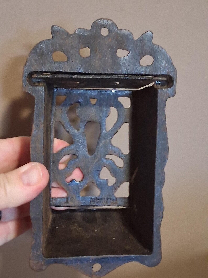 May include: A cast iron match holder with a decorative floral design. The holder is rectangular with a small shelf and a slot for matches. The metal has a dark, aged appearance, suggesting it is vintage or antique.