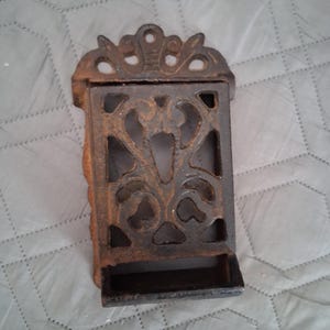 May include: A rustic, cast iron match holder with an ornate, openwork design. The rectangular holder features a decorative top and a small tray at the bottom. The metal has a dark, aged appearance, suggesting an antique or vintage style.