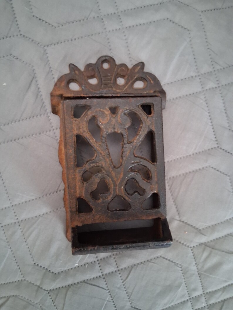 May include: A rustic, cast iron match holder with an ornate, openwork design. The rectangular holder features a decorative top and a small tray at the bottom. The metal has a dark, aged appearance, suggesting an antique or vintage style.