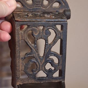 May include: An antique black cast iron match holder with a hinged lid and decorative openwork design. The holder features a small tray at the bottom and is held by a hand.