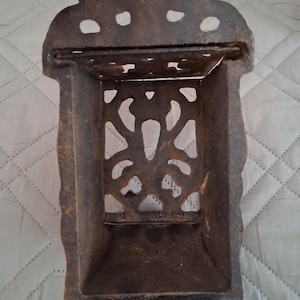 May include: A vintage, cast iron match holder with a dark brown, aged finish. The rectangular holder features an ornate, openwork design and a small hole at the top for hanging. The overall design is decorative and functional.