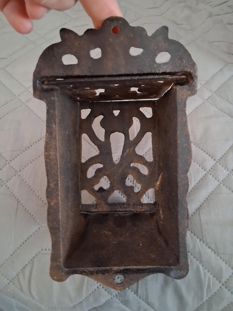 May include: A vintage, cast iron match holder with a dark brown, aged finish. The rectangular holder features an ornate, openwork design and a small hole at the top for hanging. The overall design is decorative and functional.