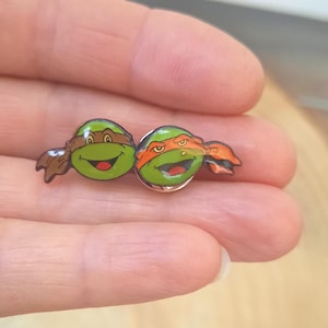 Op de afbeelding: Twee kleine ronde emaille pins met de gezichten van groene cartoon-schildpadden. De ene schildpad draagt een bruin oogmasker en kijkt glimlachend, terwijl de andere een oranje oogmasker draagt en een lachende uitdrukking heeft.