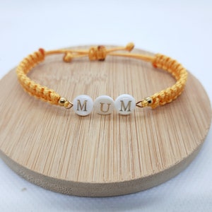 Op de afbeelding: Een gele macramé armband met het woord "MUM" in witte letterkralen. De armband heeft verstelbare schuifknopen en goudkleurige accenten. De armband wordt gepresenteerd op een houten oppervlak.