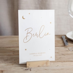 May include: A white card with gold script that reads "Berlin" with the names "Amber & Marcus" and the date "26th April 2024" below. The card is decorated with gold stars and a crescent moon. It is displayed on a wooden stand.