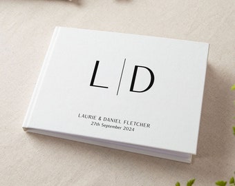 以下が含まれることがあります： 「L」と「D」のイニシャルが垂直線で区切られた白いゲストブック。「Laurie & Daniel Fletcher 27th September 2024」というテキストがイニシャルの下に印刷されています。