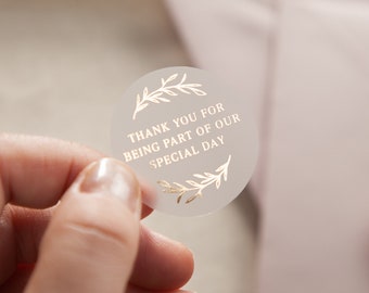Op de afbeelding: Ronde, transparante sticker met gouden folie-lettering met de tekst "Bedankt dat je deel uitmaakte van onze speciale dag". De sticker heeft een gouden folie-bladontwerp rond de tekst.