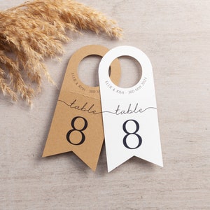 May include: Two brown and white table number cards with a hole at the top for hanging. The cards are shaped like a banner and have the number 8 printed on them. The text "table" is printed above the number. The text "Ellie & Josh - 3rd May 2021" is printed on the top of the cards.