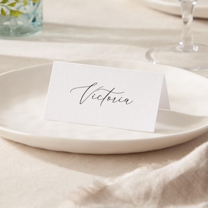 May include: White place card with the name "Victoria" written in black cursive script.