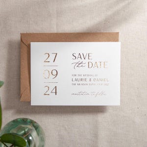 May include: A white save the date card with gold foil lettering. The date is 27th September 2024. The text reads "Save the Date" for the wedding of Laurie & Daniel. The Meadow Barn, CV37 0GE. Invitation to follow.