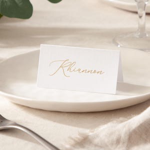 May include: A white place card with the name "Rhiannon" in gold script is displayed on a white plate. The card is folded and stands upright, suggesting a wedding or event setting. The background includes a fork and a linen tablecloth.