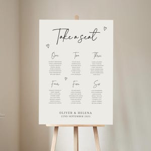 May include: A white wedding seating chart with the text "Take a seat" in black cursive script. The chart is divided into six columns labelled "One", "Two", "Three", "Four", "Five", and "Six". Each column lists names of wedding guests. The bottom of the chart includes the names "Oliver & Helena" and the date "22nd September 2025".
