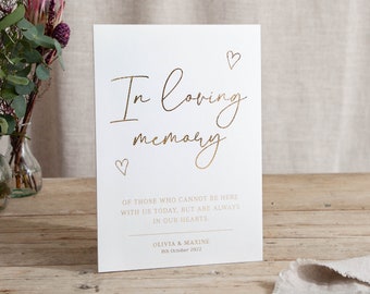 May include: A white card with gold lettering that reads "In loving memory" with two small hearts above the text and two small hearts below the text. The card also includes the text "Of those who cannot be here with us today, but are always in our hearts." and "Olivia & Maxine 8th October 2022".