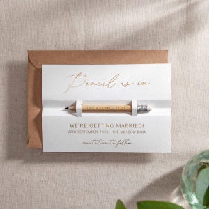 May include: A white card with gold lettering that says "Pencil us in" and "We're getting married!" with a date and location. A small wooden pencil is sticking out of the card.