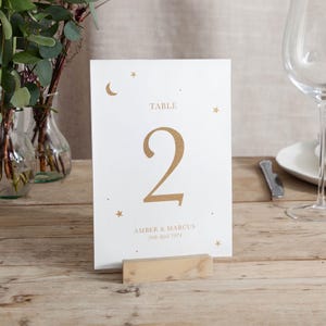 May include: A white table card with gold text and a wooden base. The card features the word "TABLE", the number "2", and the names "AMBER & MARCUS" with the date "26th April 2024". Gold stars and a crescent moon decorate the top.