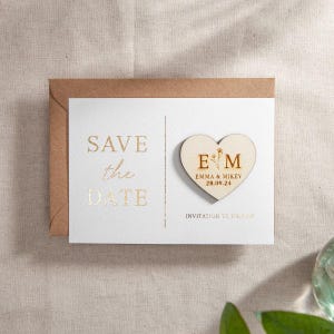 May include: A wedding announcement card with a wooden heart magnet. The card reads "Save the Date" in gold lettering. The heart magnet has the initials "E M" and the date "28.09.24". The card is in a brown envelope.