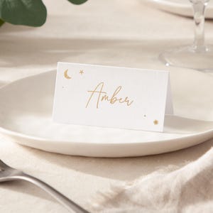 May include: White place card with gold foil lettering and a crescent moon and stars design. The name "Amber" is written in cursive.