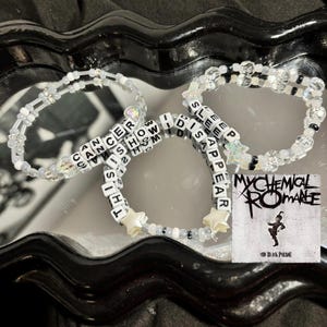 May include: Three white beaded bracelets with black letter beads spelling out the phrases "Cancer", "This is how", and "I disappear" with a white star charm on each bracelet. The bracelets are on a black surface with a My Chemical Romance album cover in the background.