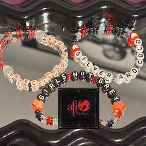 Op de afbeelding: Drie armbanden met kralen in wit, rood, zwart en zilver. Eén armband spelt "THIS CELLUL DREAM", een andere "SILVER AND COLD" en de derde "GIRLS NOT GREY". Er is ook een zwart vierkant met tekst en een logo.