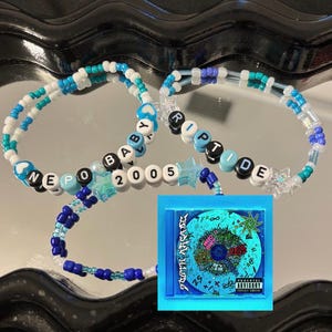 May include: Three beaded bracelets with white, blue, and teal beads spelling out "NEPO BABY 2005" and "RIPTIDE". A CD case with a blue and green graphic design is also present.
