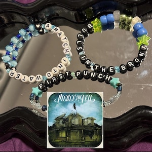 May include: Three beaded bracelets with blue and green beads and white letters spelling out "A Match Into First Punch In The Bronx". The bracelets are on a mirrored surface with a CD for the album "Collide With The Sky" by Pierce The Veil.